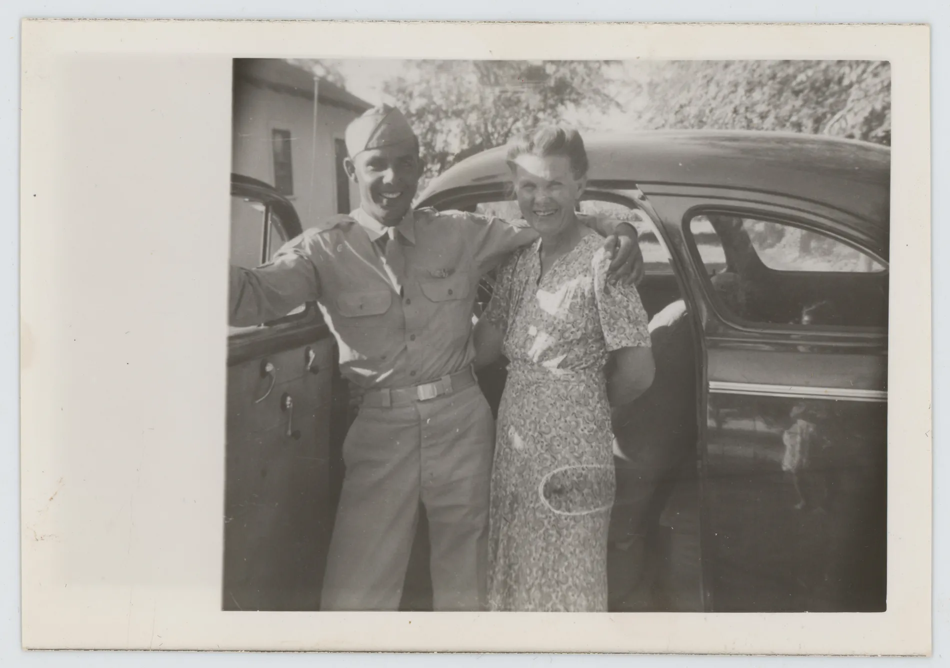 This photograph depicts Oscar Allison standing with his mother, Ollie, next to a car. This photograph appears to be from before Oscar's time as a prisoner of war. A distorted reflection of the photographer can be seen on the car.