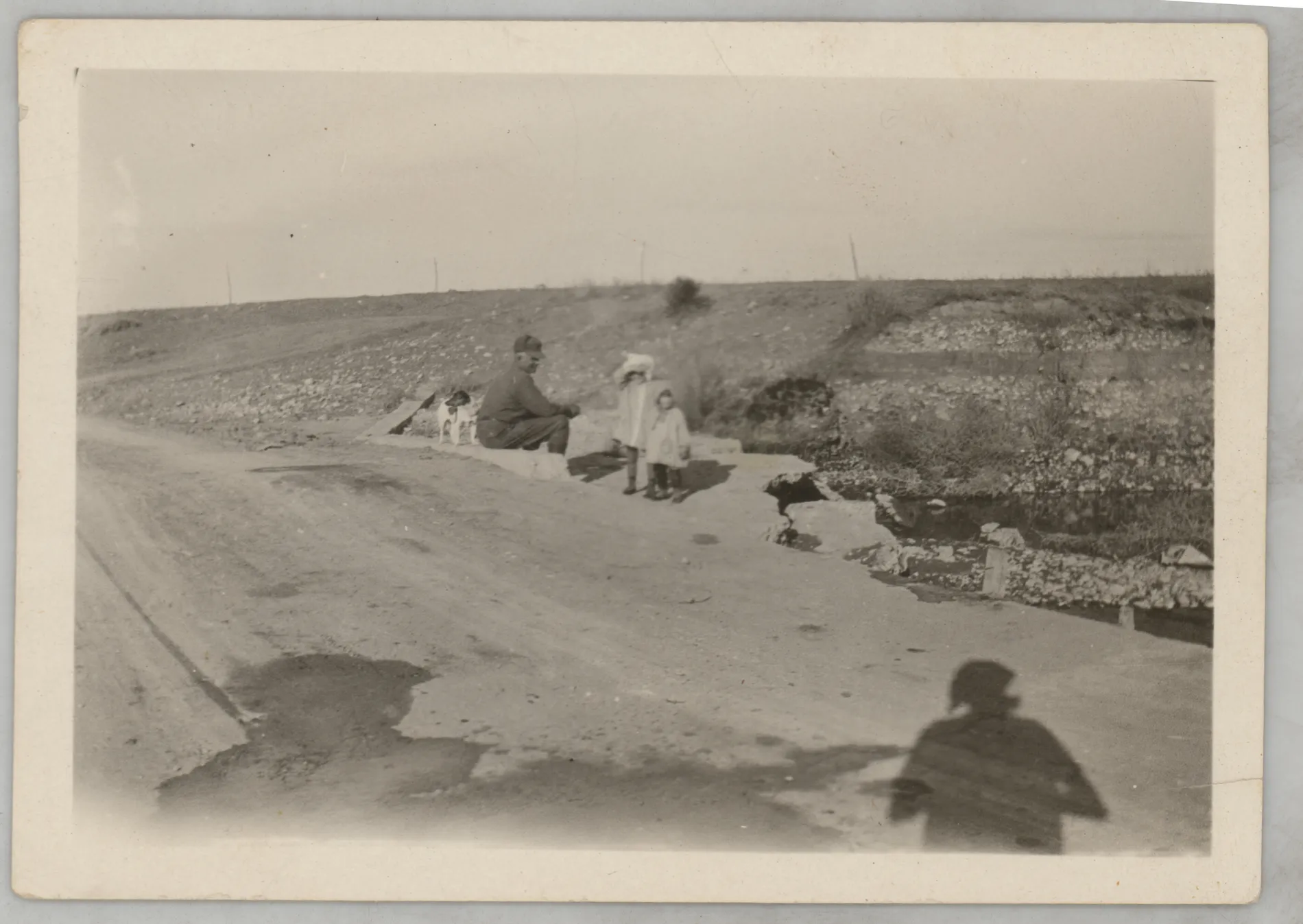 This photograph depicts Sam, Sammy, and Jean McKinstry standing and sitting on a road next to the Felix River. A dog is to the left of Sam.