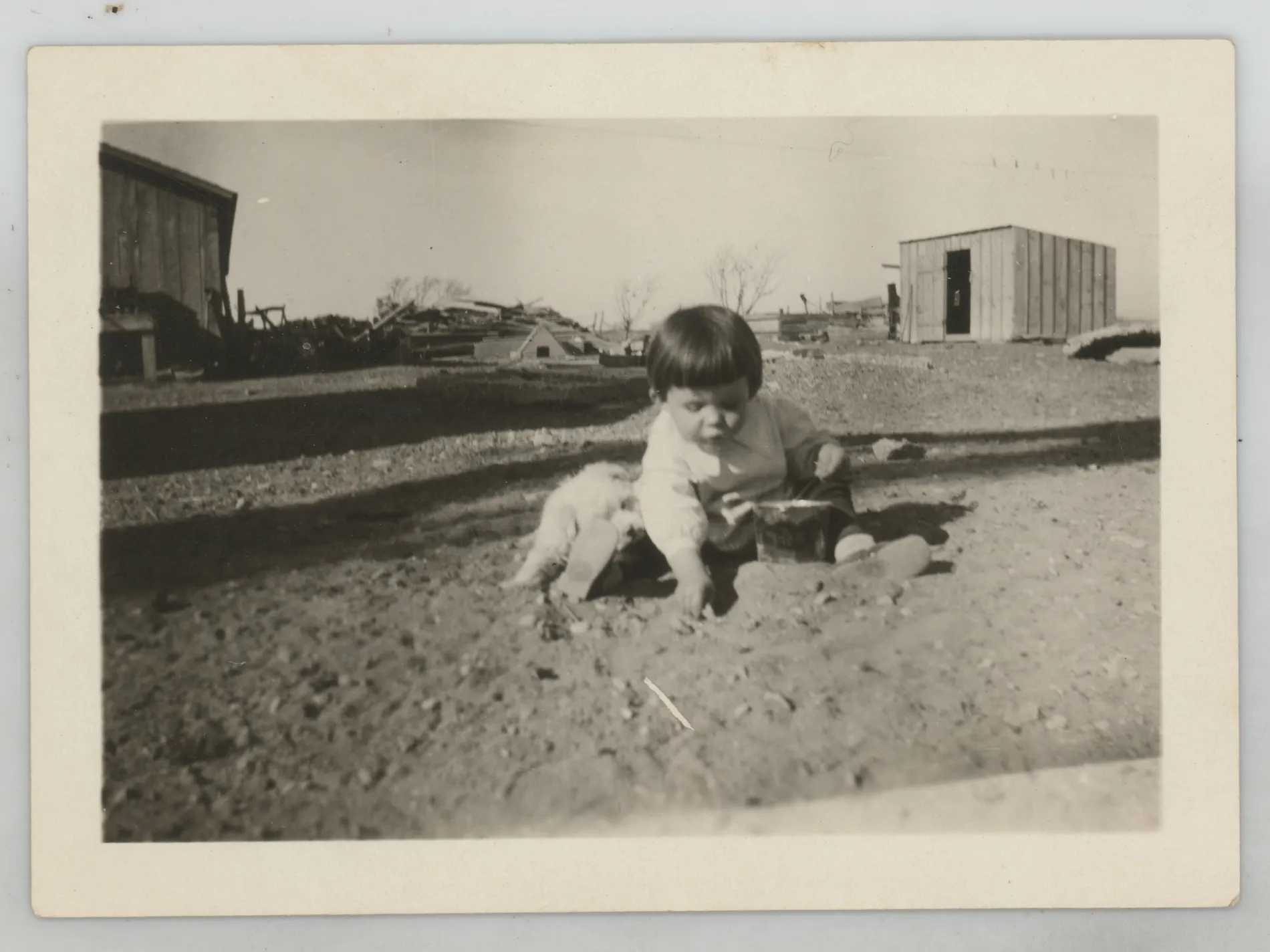 This photograph depicts Sammy McKinstry playing on the ground in the dirt with a dog near by. Two structures are in the background.