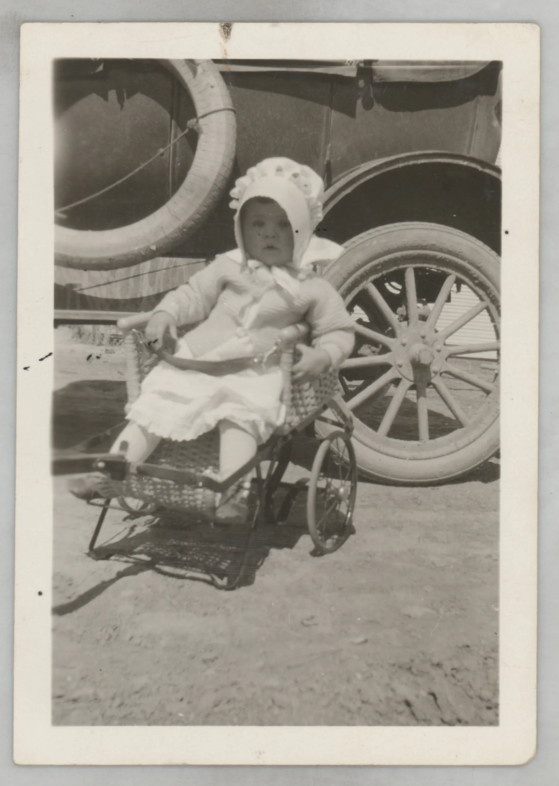 This photograph depicts Sammy McKinstry in a whicker stroller or chair on wheels. She is next a car tire in this photograph.