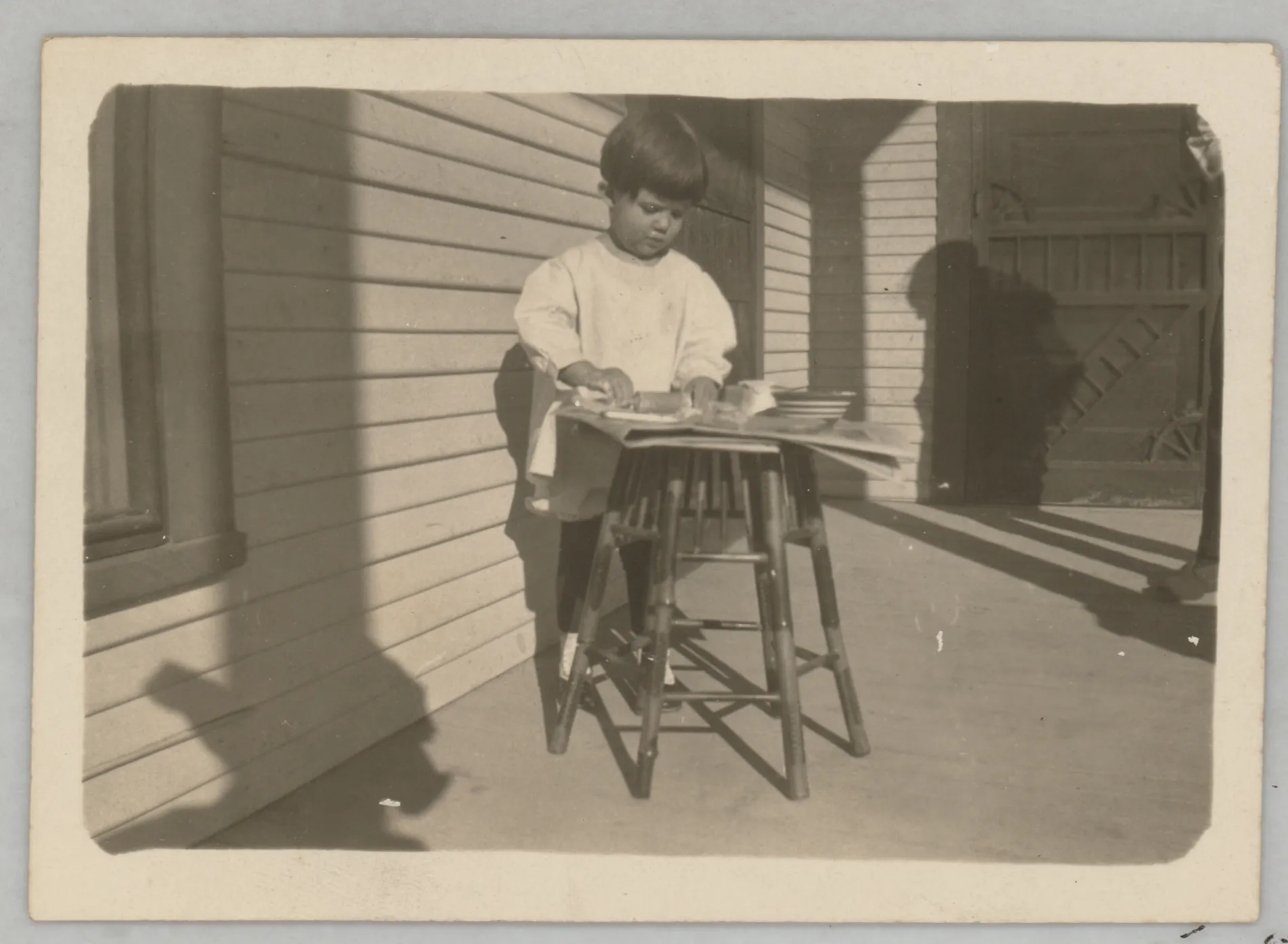 This photograph depicts Sammy McKinstry using miniature cooking implements to pretend to cook. She is standing on the porch of a house. An unknown man casts a shadow on the right. This man's shoes match those of Austin Swann.
