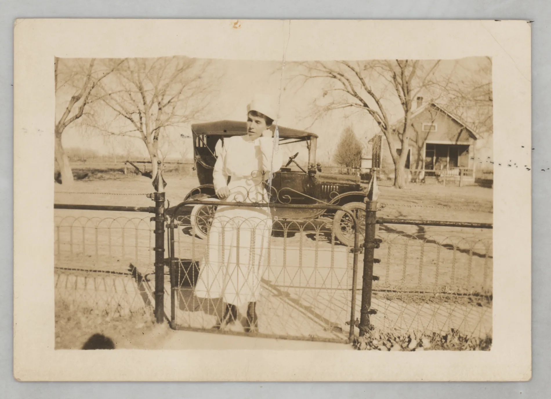 This photograph depicts an unknown woman in the garb of a nurse about to enter through a gate. A car is in the background.