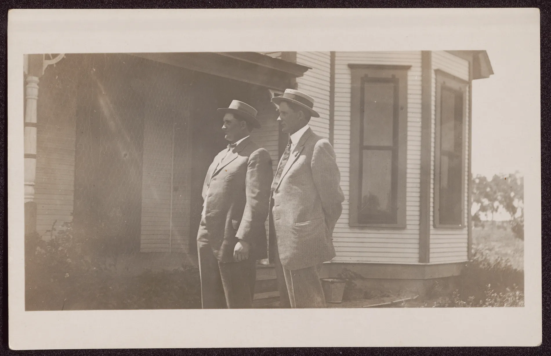 This photograph depicts Jim and Sam McKinstry standing next to a house in formal wear and hats. It is possible that this photograph is from November 7, 1912, the date that brothers Jim and Sam McKinstry married sisters Minnie and Loveta Swann.