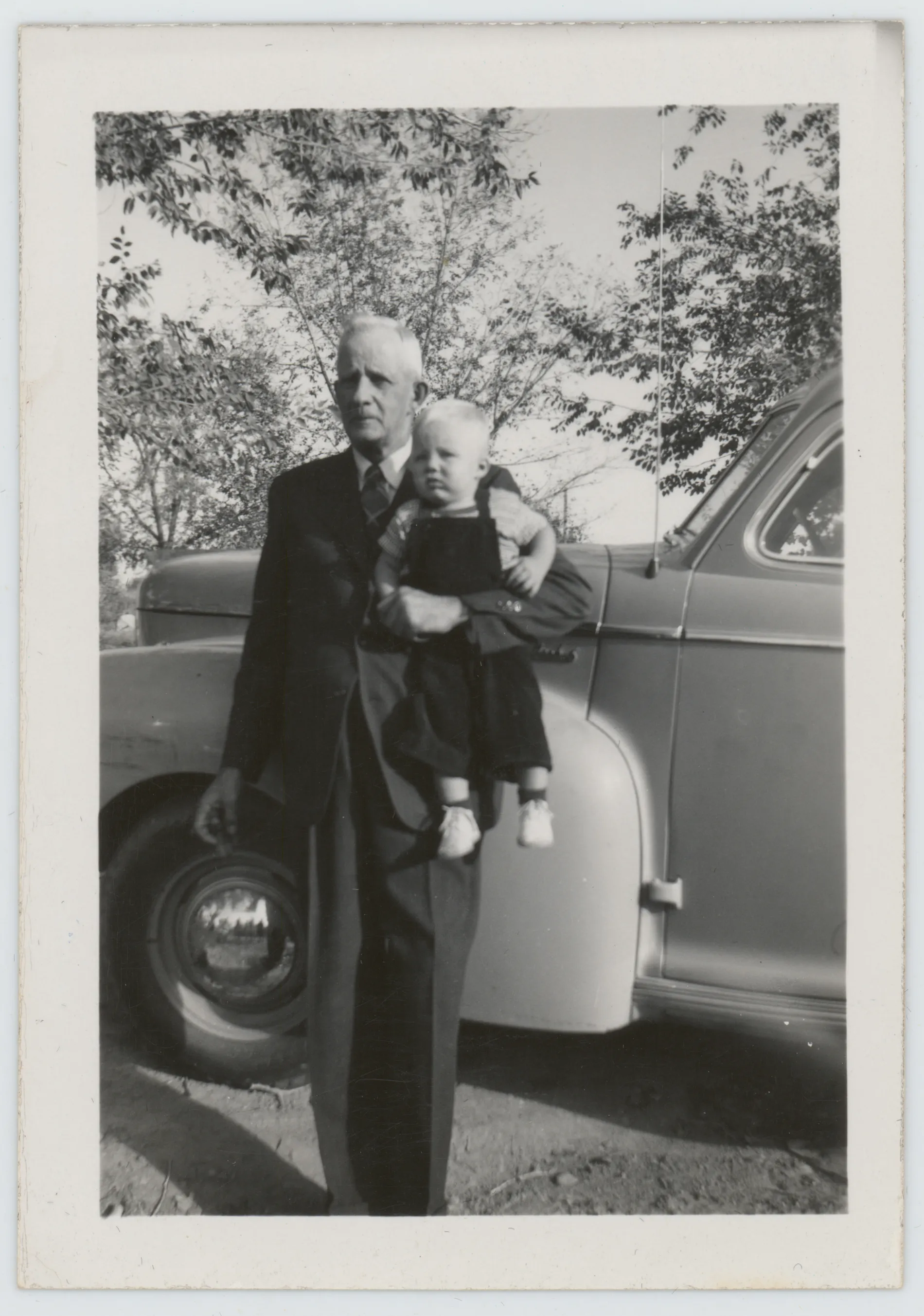 This photograph depicts Sam McKinstry holding his grandson, Mack. They are next to a car.