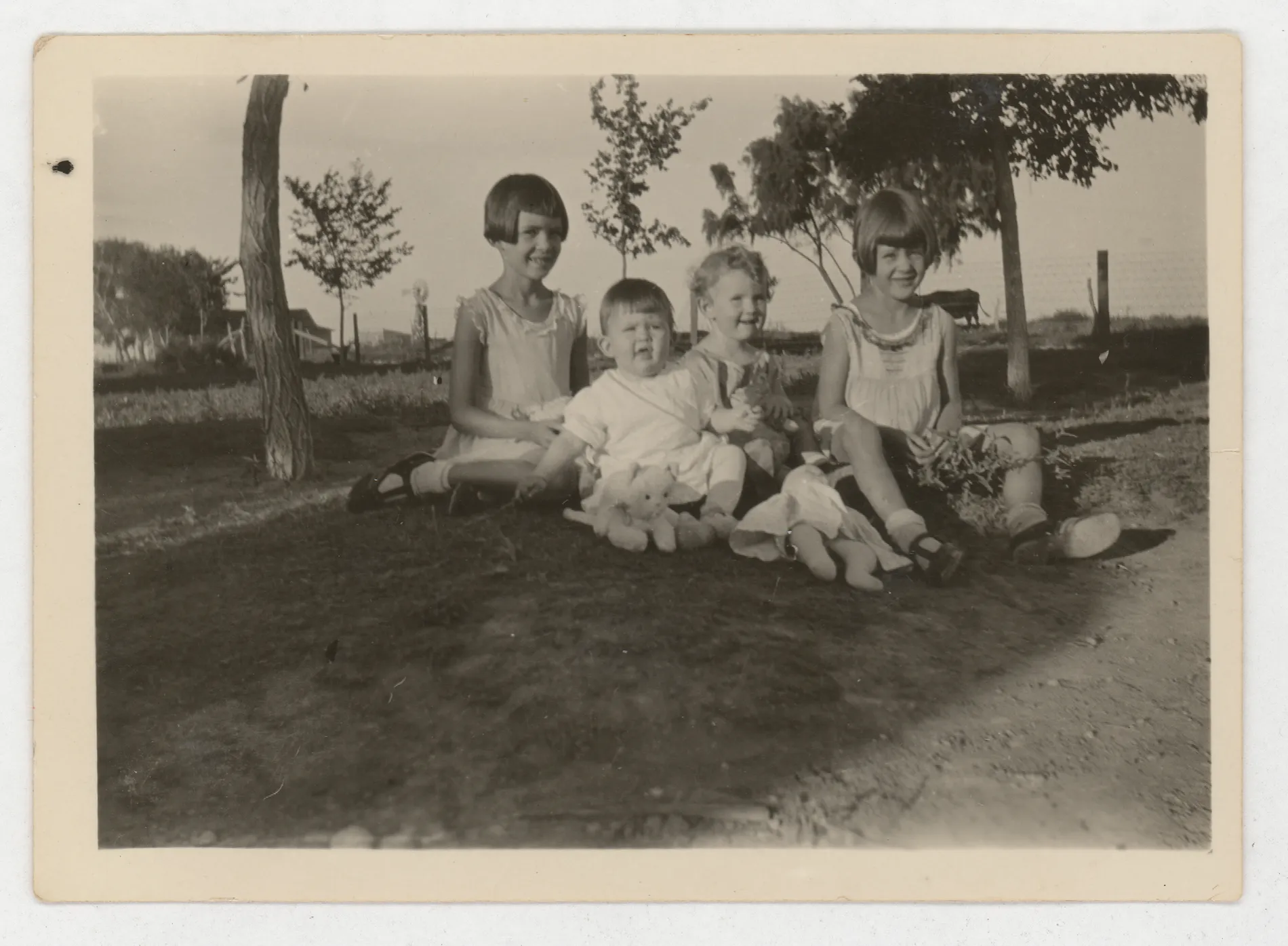 This photograph depicts Sammy, Mildred, Peggy, and Jean McKinstry sitting on the amidst some trees.