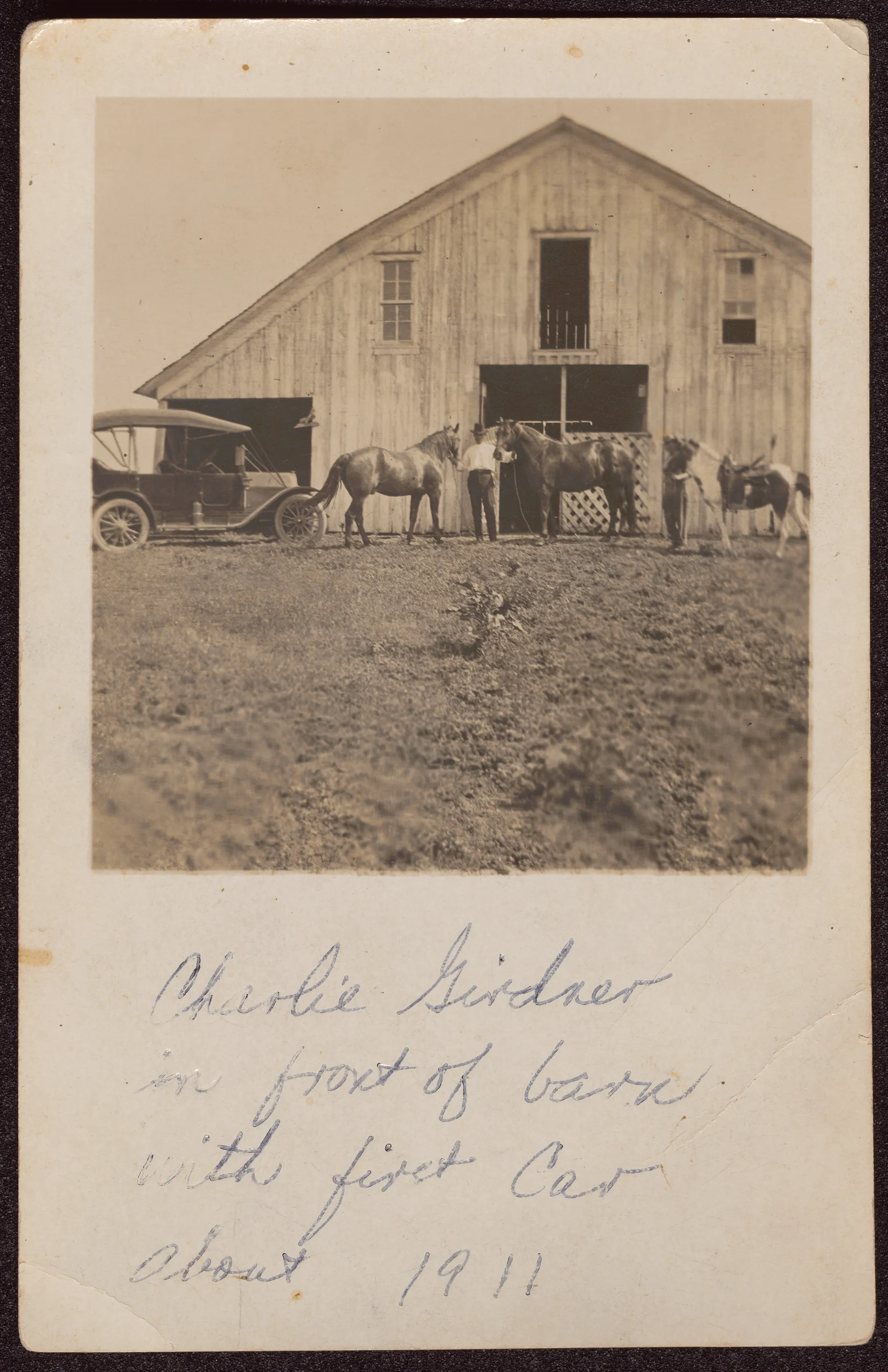 This photograph depicts Charlie Girdner standing in front of a barn. A car is to the left. Three horses are also visible. There is another figure behind a horse. The verso identifies them as "Jute (Doc) Girdner.