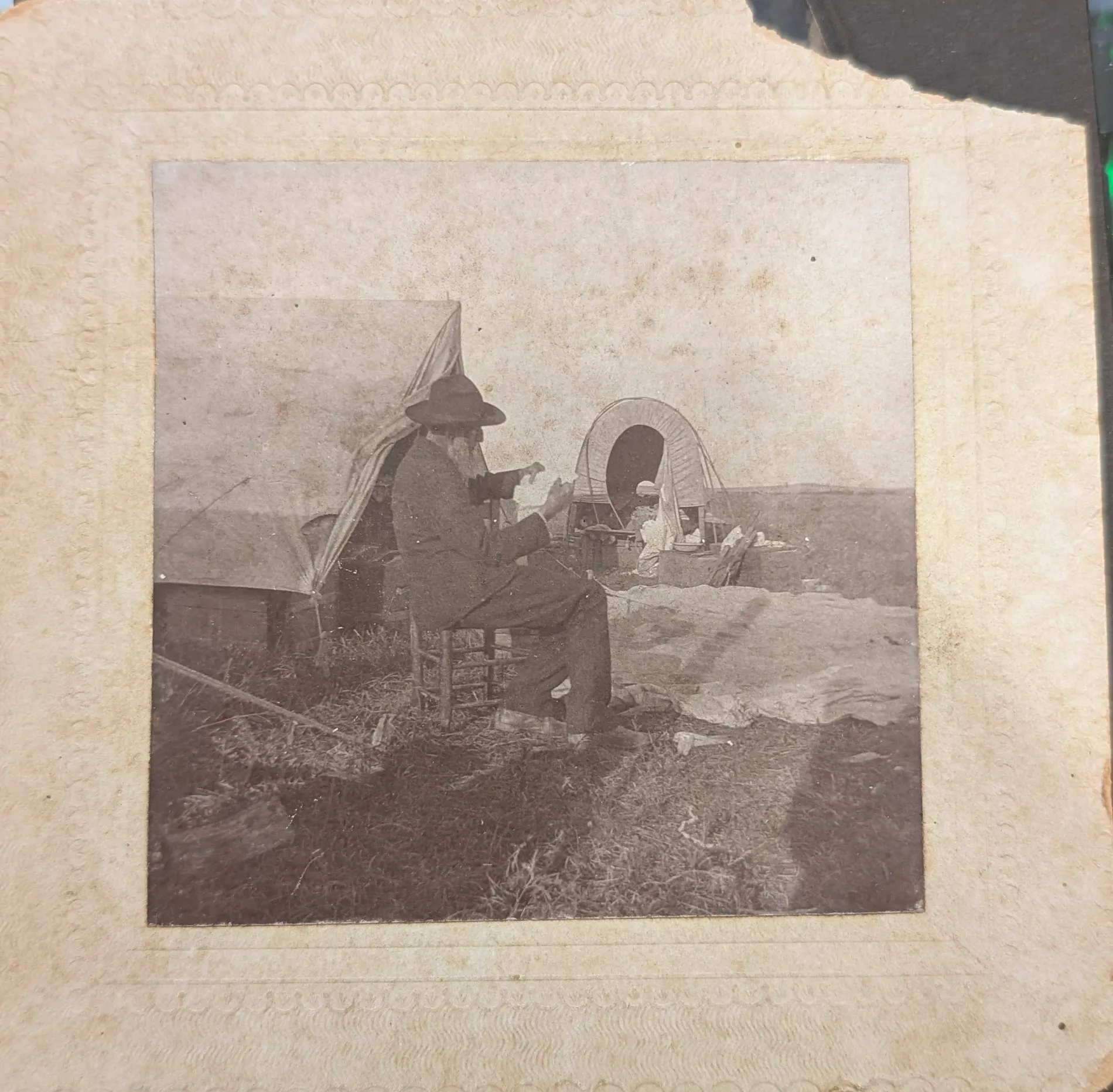 An elderly man with a full white beard sits in profile on a ladder-back chair in a camp on open prairie. He wears a dark coat and trousers and a broad-brimmed hat, and holds his hands up in front of him around something small that cannot be made out. Behind him a canvas wall tent stands with its flap thrown back. To the right a covered wagon is drawn up with its bows and cover in place; a figure in light-coloured clothing stands at the tailgate among boxes and gear, and canvas or bedding is spread on the ground beside it. Cut grass, scattered sticks and a low fence rail lie in the foreground; the horizon is flat and empty. The print is roughly square, mounted on a cream card with a blind-embossed scalloped border inside two ruled lines; the mount is torn away at the upper right corner.