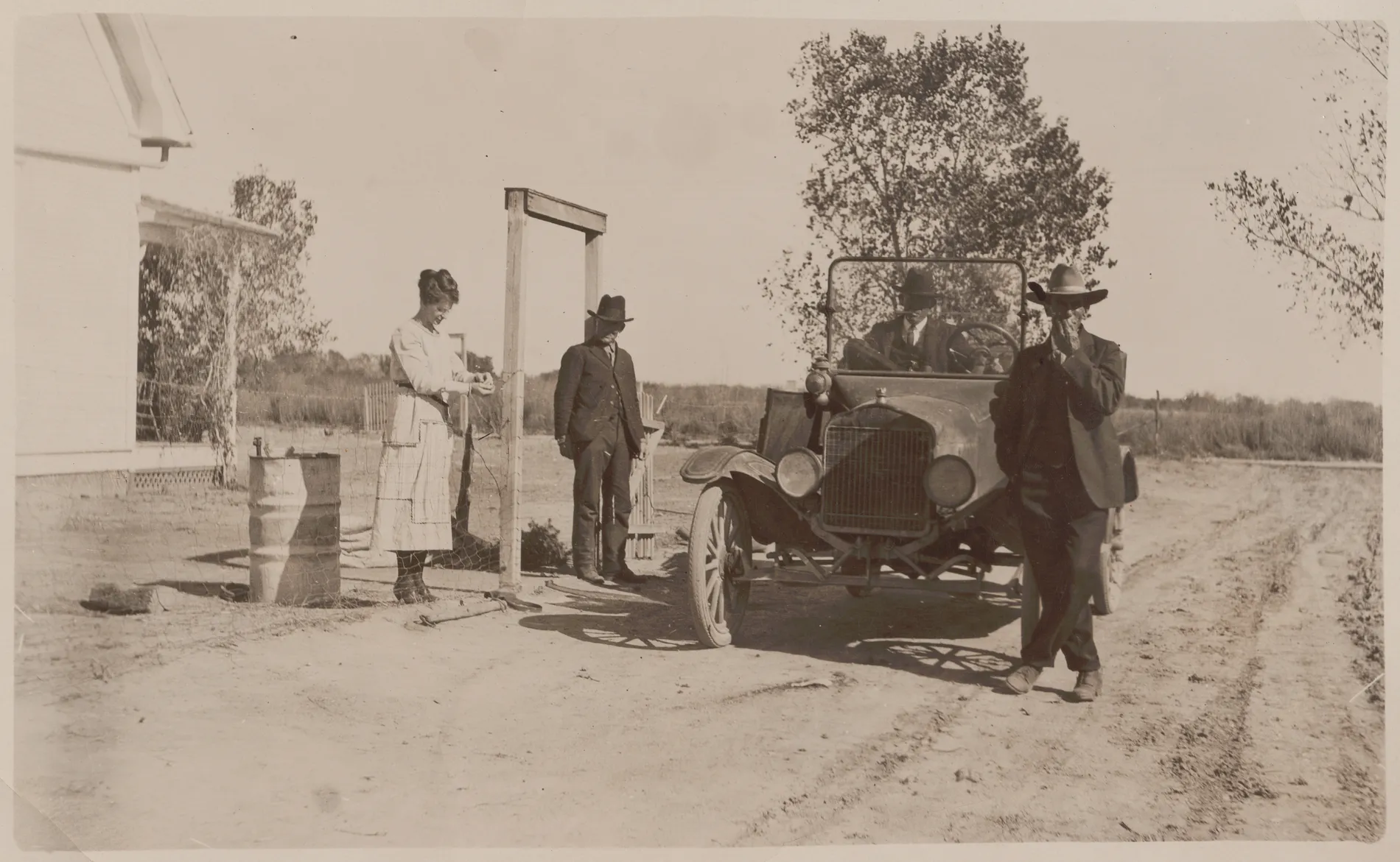 This photograph depicts Austin Swann sitting in a car, Eva Swann standing in the gate, a Mr. Will Lane next to the gate, and Mr. Fay Irwin standing in front of the car.