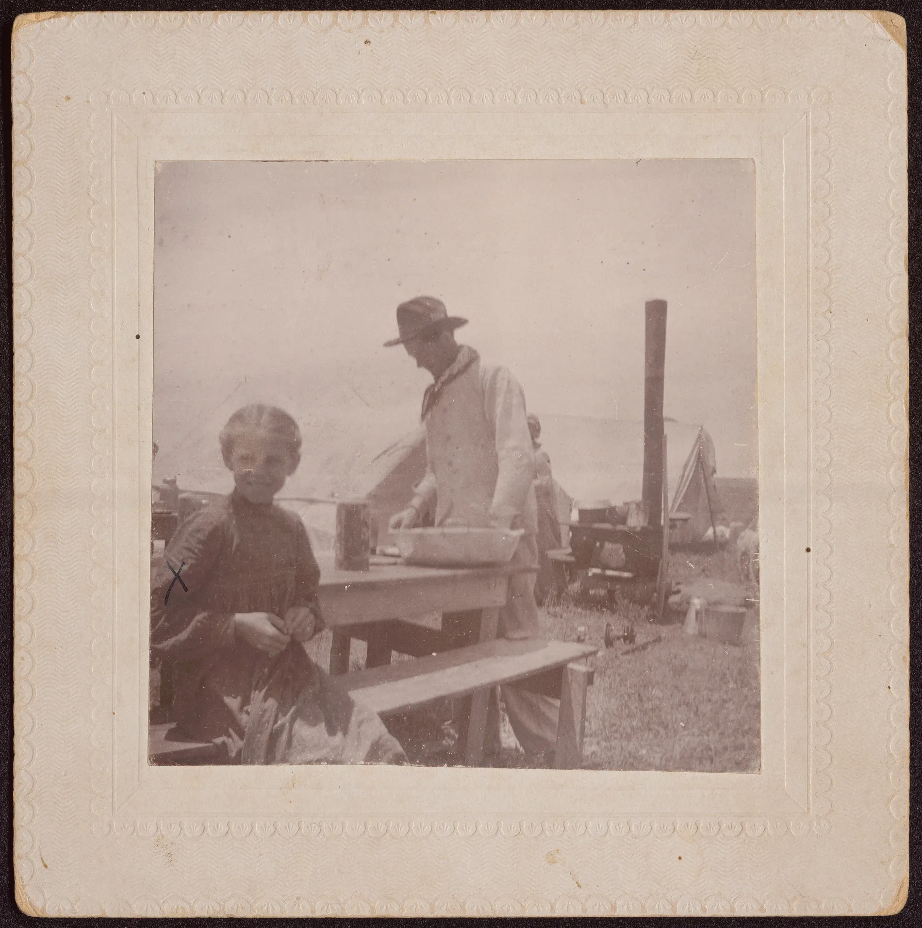 This photograph depicts Loveta Swann (marked with an X) sitting down on a bench with cook Pete Lovall in the background. Typical cooking equipment is in view.